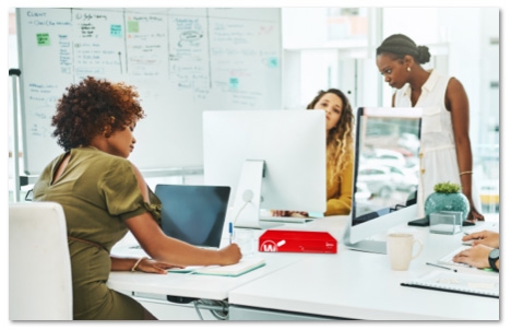 3 women working in a bright office with a red WatchGuard tabletop Firebox on the desk