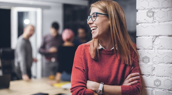 Lächelnde Frau mit blonden Haaren, grüner Brille und einem orangefarbenen Pullover lehnt an einer weißen Ziegelwand.